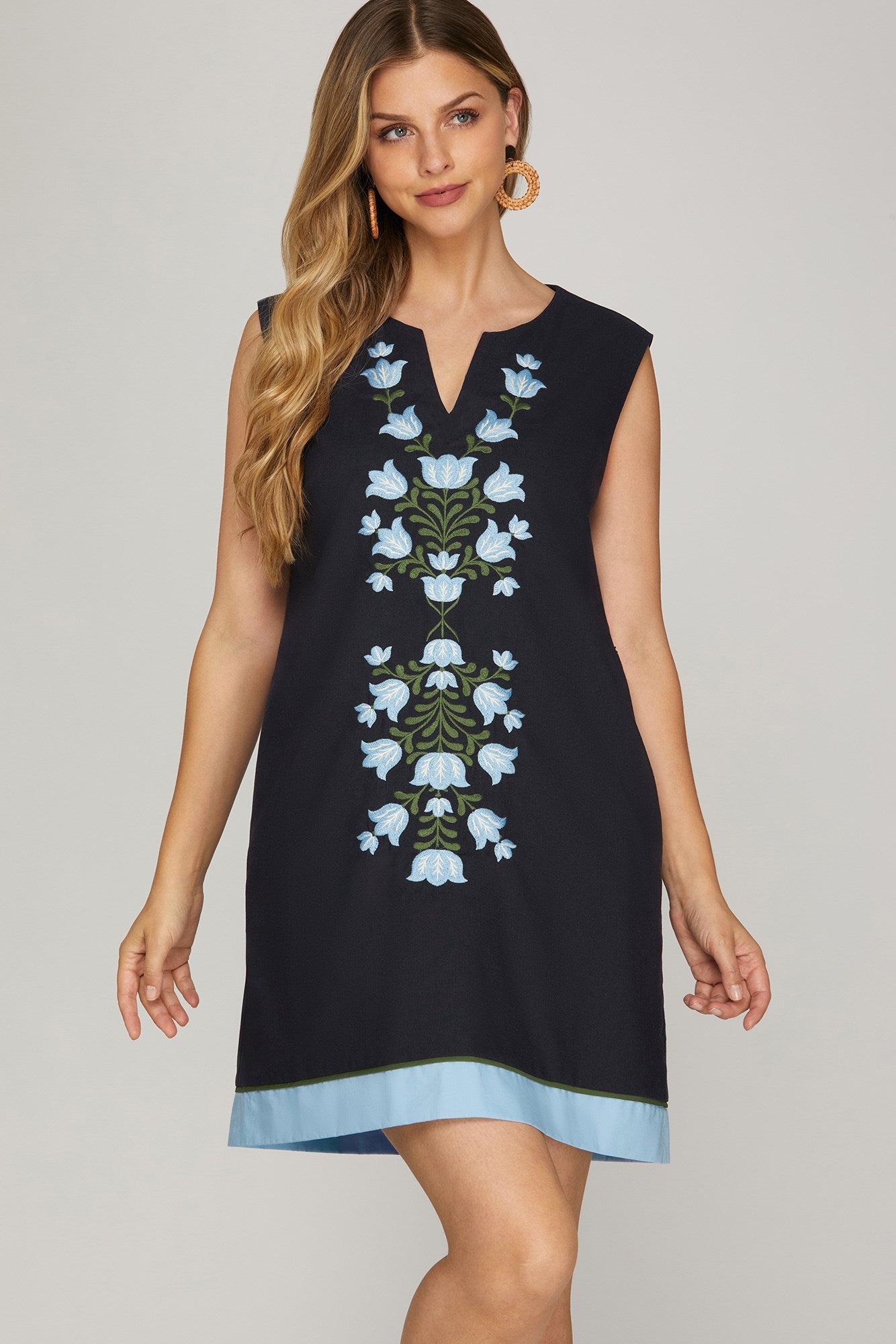 Woman wearing navy sleeveless mini shift dress with blue and white floral embroidery down the front, contrast sky-blue hem, and side pockets